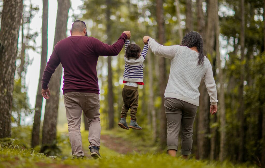 A family is walking together in a forested area, seen from behind. The parents, each holding one of their child's hands, lift the child joyfully into the air. The father, wearing a burgundy sweater and beige pants, and the mother, dressed in a white sweater and green pants, walk side by side along a path lined with tall trees. The child, in a striped sweater and brown pants, kicks their legs in excitement. The lush greenery and towering trees create a serene, nature-filled backdrop, capturing a playful and heartwarming family moment in the outdoors.