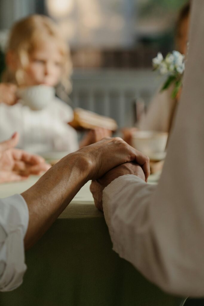 An intimate moment is captured as two elderly individuals hold hands across a table. Their hands are gently clasped, symbolizing affection and support. The soft lighting gives a sense of warmth and tenderness. In the background, a young child with light blonde hair can be seen drinking from a cup, but the focus remains on the joined hands in the foreground. The blurred background adds to the feeling of quiet connection, making the scene feel calm and reflective. The overall setting hints at a family gathering or shared meal, evoking a sense of generational love and togetherness.