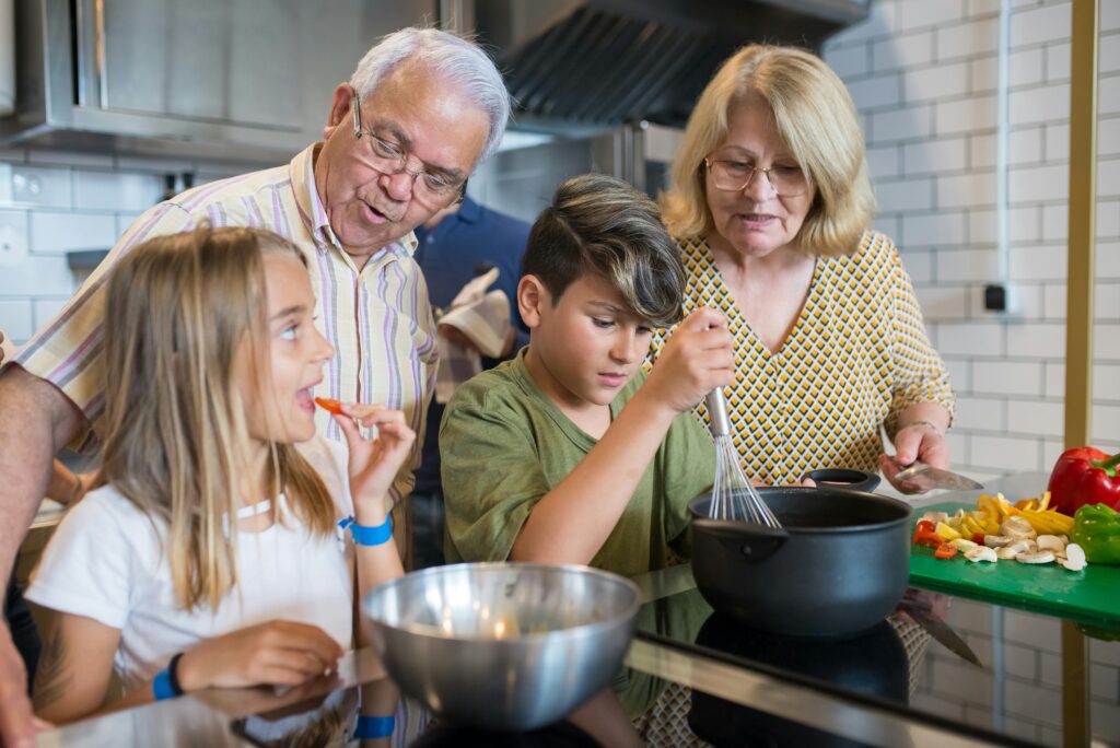 A cheerful family moment unfolds in the kitchen, with grandparents and grandchildren cooking together. The grandfather, with gray hair and glasses, watches closely as the young boy, focused, whisks ingredients in a pot. The grandmother, also with gray hair, stands nearby, assisting and cutting vegetables on a green cutting board. The young girl, smiling, enjoys a slice of bell pepper as a snack while observing the cooking process. The scene is filled with fresh vegetables and cooking tools, creating a warm and lively atmosphere as the family bonds over preparing a meal together.