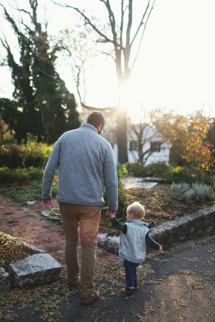 A man and a young child are walking hand-in-hand down a peaceful path in an outdoor setting. Both are dressed warmly, with the man wearing a gray sweater and the child in a gray vest over a plaid shirt. The scene is backlit by the soft glow of the setting sun, casting a warm, golden light over the surroundings. Fallen leaves scatter the path, and the trees and plants in the background add to the calm, autumnal atmosphere. The moment feels serene and full of tenderness as the man gently leads the child on their walk.