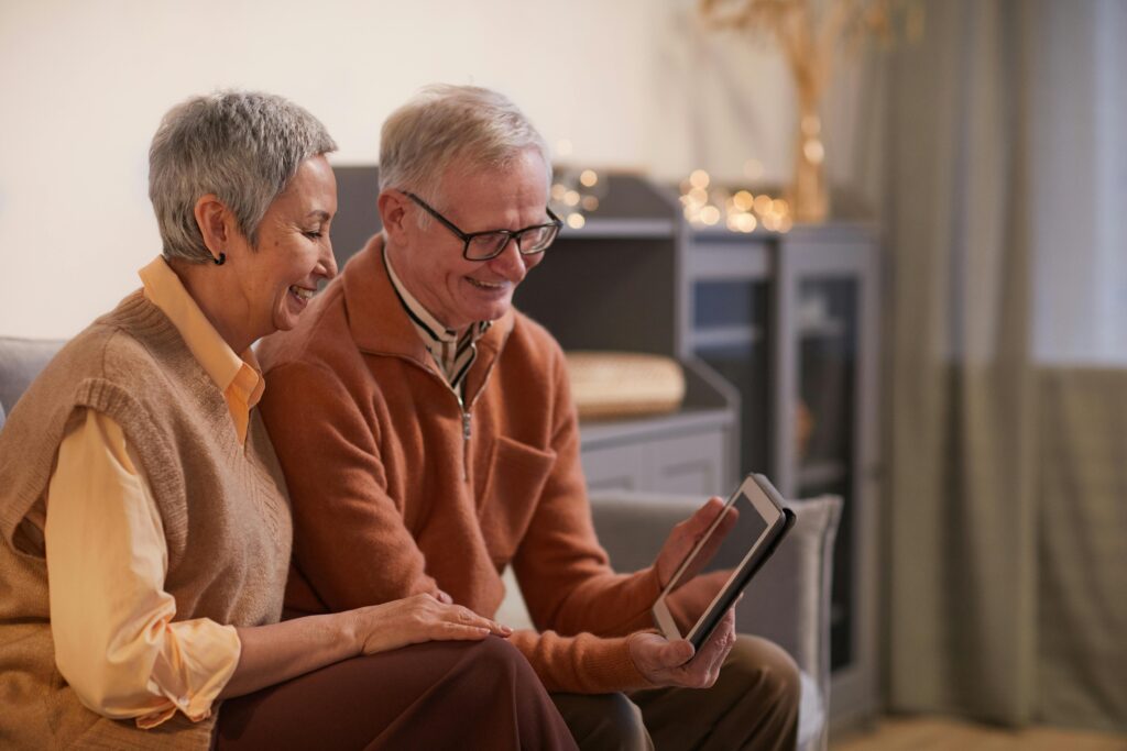 An elderly couple sits together on a cozy couch, both smiling as they look at a tablet. The woman, with short gray hair, wears a beige sweater vest over a yellow blouse, while the man, with light-colored hair and glasses, is dressed in an orange zip-up sweater. Their posture reflects comfort and companionship as they share a warm, lighthearted moment. The soft lighting and simple decor in the background, including shelves and glowing string lights, add to the inviting and peaceful atmosphere of their home setting.