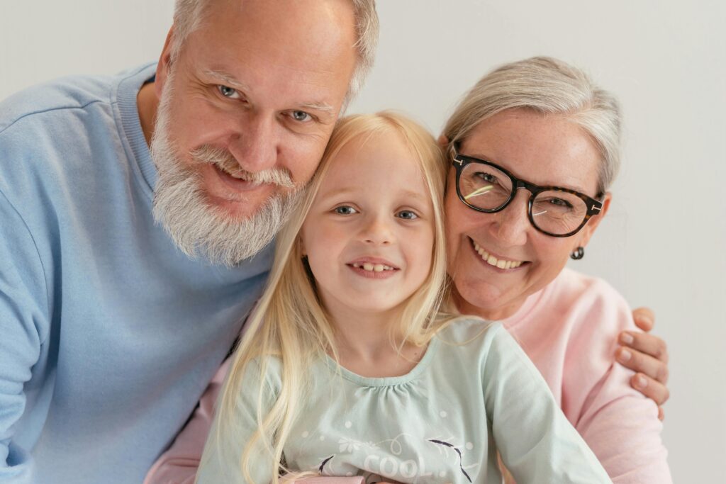 A smiling elderly couple poses with their granddaughter, creating a heartwarming family portrait. The grandfather, with a white beard and a blue sweater, leans in closely, while the grandmother, wearing glasses and a pink sweater, wraps her arm around the child. The young girl, with long blonde hair and a soft teal shirt, smiles sweetly at the camera. The bright, simple background keeps the focus on the joyful expressions and close bond between the three. The portrait exudes love, warmth, and familial connection.