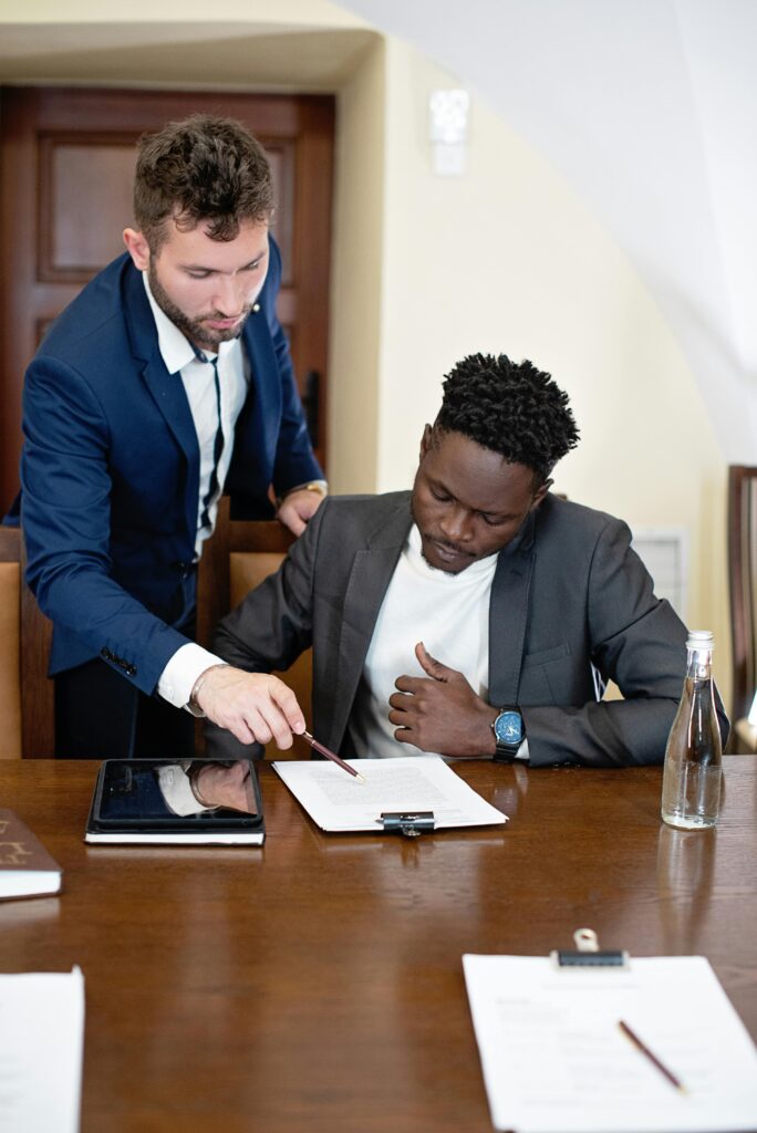 Two men in business attire are working together at a table. The man standing, dressed in a navy suit, points at a document with a pen, explaining something to the man seated, who is focused on the paper in front of him. The seated man, wearing a dark gray suit and white shirt, listens intently, showing a thoughtful expression as he follows along. On the table, there are papers, a tablet, and a bottle of water, suggesting a professional meeting or consultation. The setting has a formal, business-like atmosphere, with the men engaged in a focused and collaborative discussion.